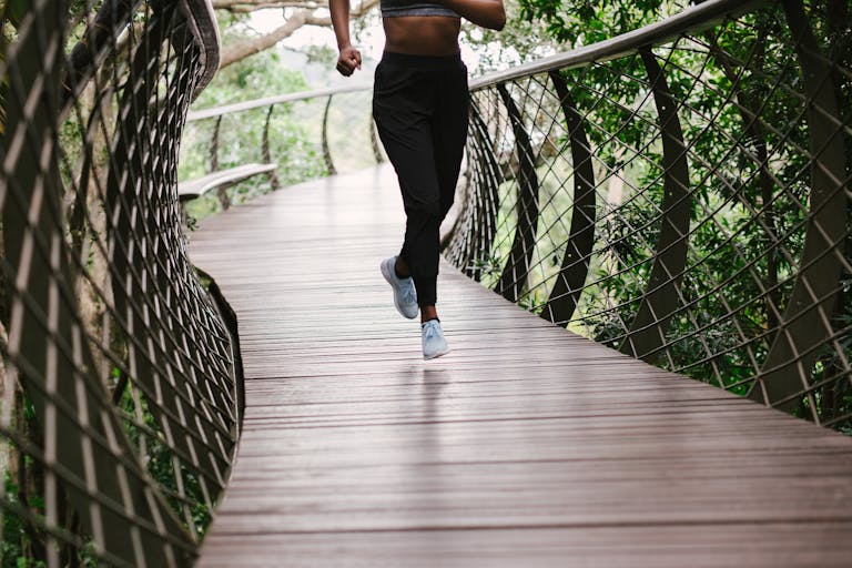 An athletic woman jogging on a nature trail bridge surrounded by greenery.